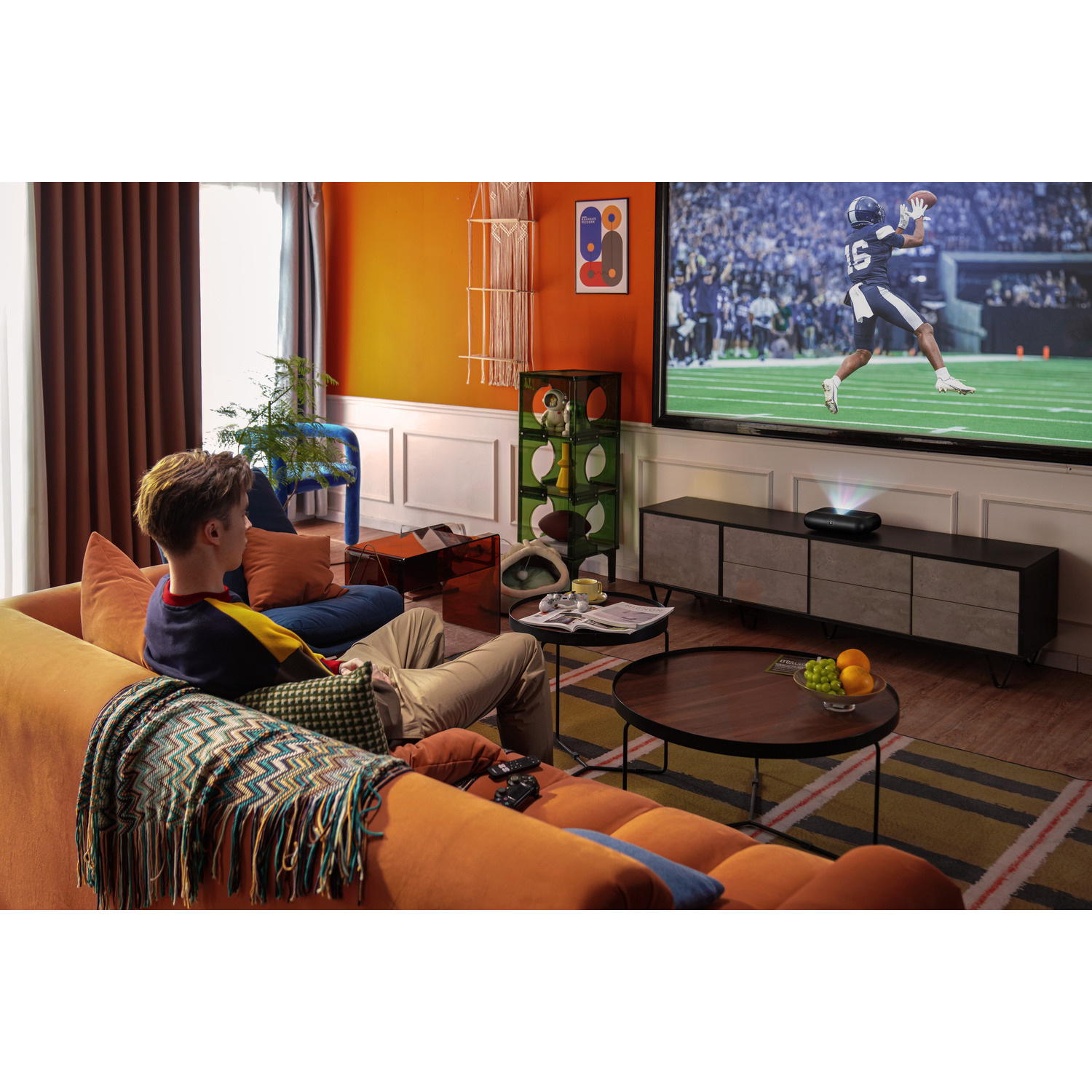Een man kijkt tv met een footballwedstrijd. De kamer heeft oranje muren en een bank.