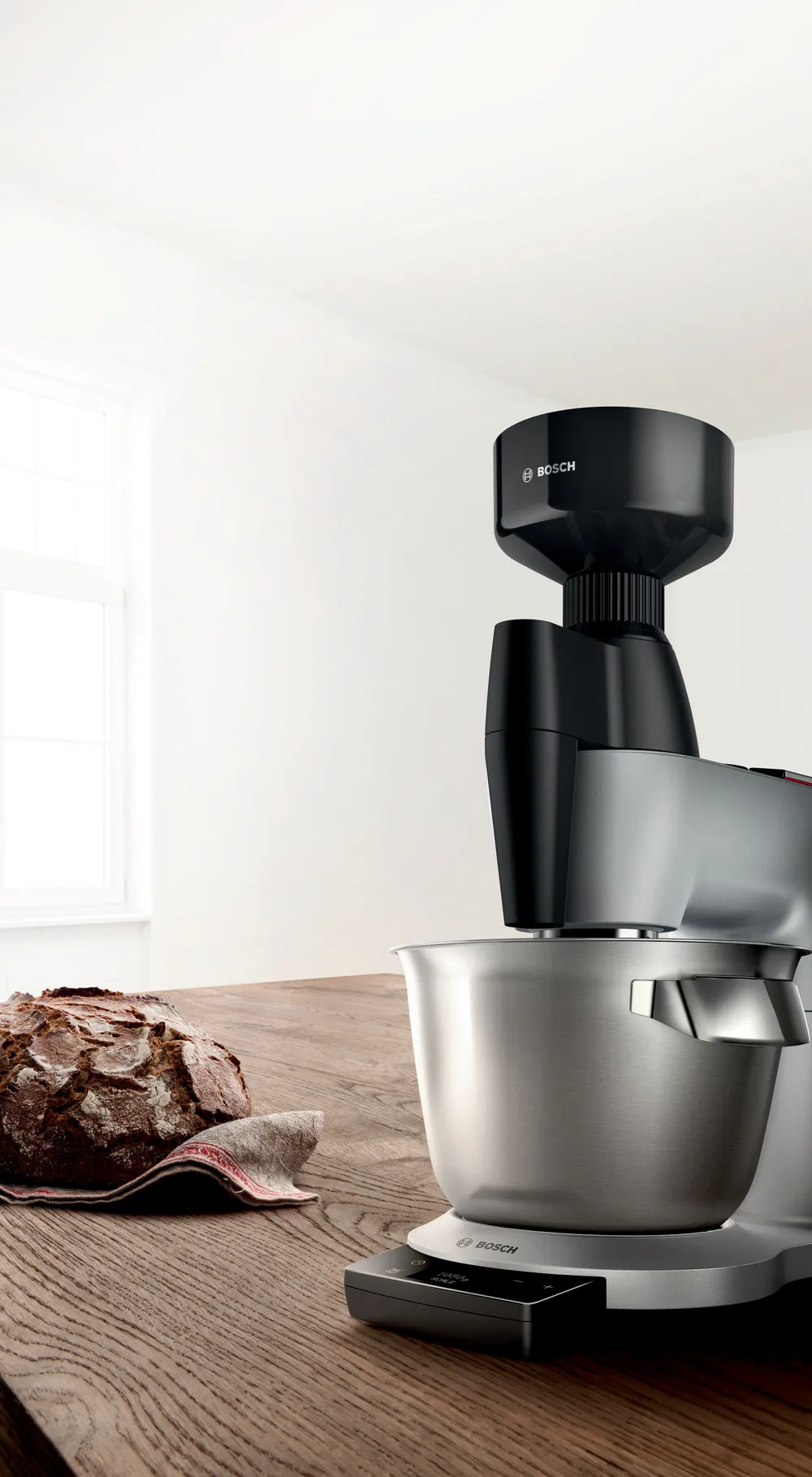 Egy Bosch konyhai mixer egy kenyérrel egy faasztalon. Fehér fal és ablak a háttérben. Fekete és ezüst színű mixer.