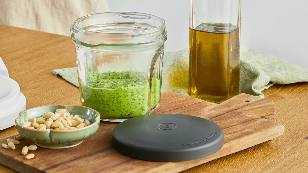 Pesto hozzávalók fa deszkán: pesto üveg, olívaolaj, fenyőmag és fedő. Háttér: világos színű kendő.