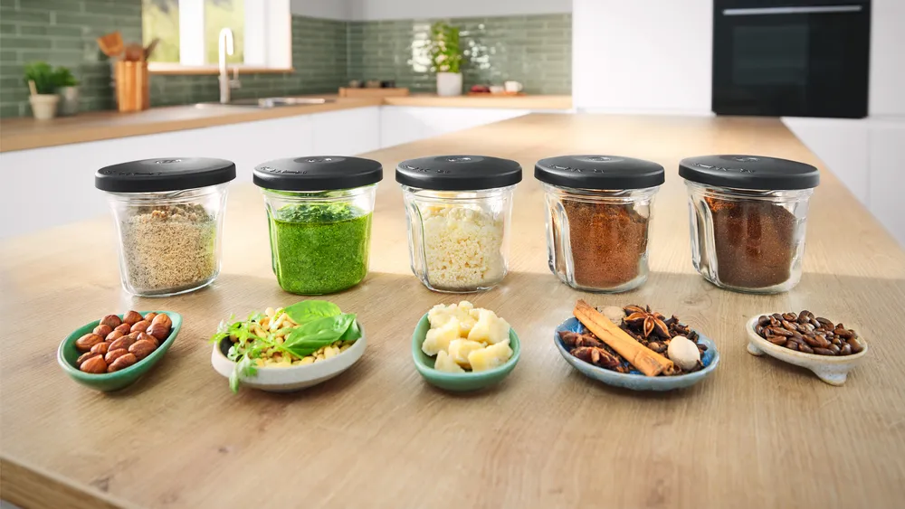 Üveg edények fűszerekkel és gyógynövényekkel, diós, fűszeres és kávébabos tálak fa felületen. Konyhai háttér.