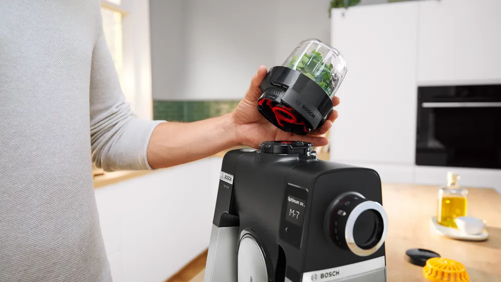 Egy kéz egy zöldfűszerekkel teli turmixpoharat helyez egy fekete Bosch konyhai robotgépre. Konyhai jelenet fehér szekrényekkel és fa pulttal.