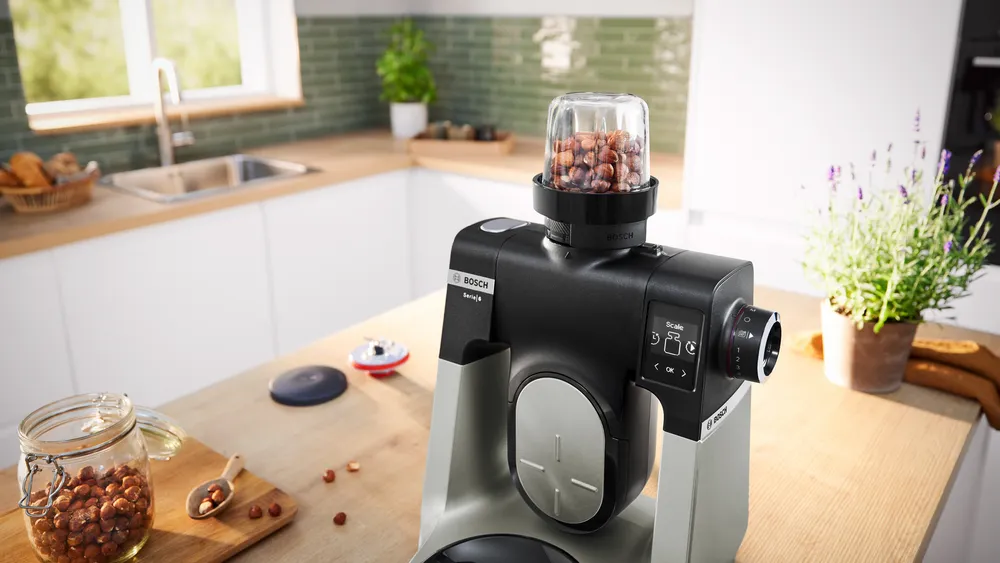 Egy Bosch konyhai robotgép dióval egy üvegtartóban. Egy üveg dióval és egy fakanál az asztalon.