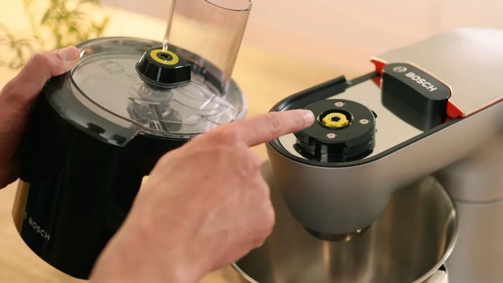 Kézben egy fekete konyhai robotgép alkatrész és egy ezüst keverő. Ujj a keverő rögzítési pontjára mutat. A "BOSCH" logó látható.