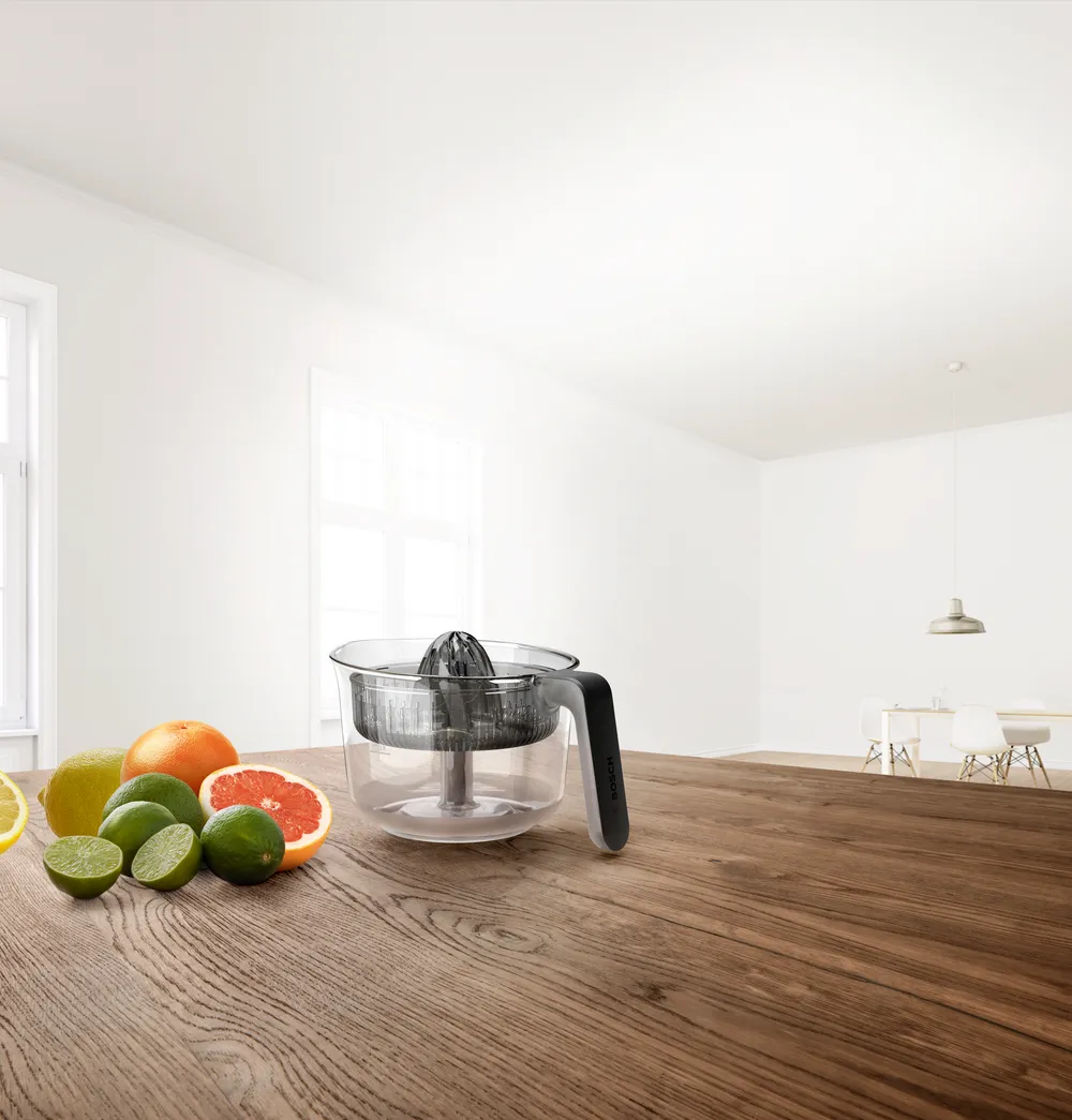 Egy gyümölcscentrifuga és citrusfélék egy faasztalon. Szoba fehér falakkal és ablakokkal. Asztalok és székek a háttérben.