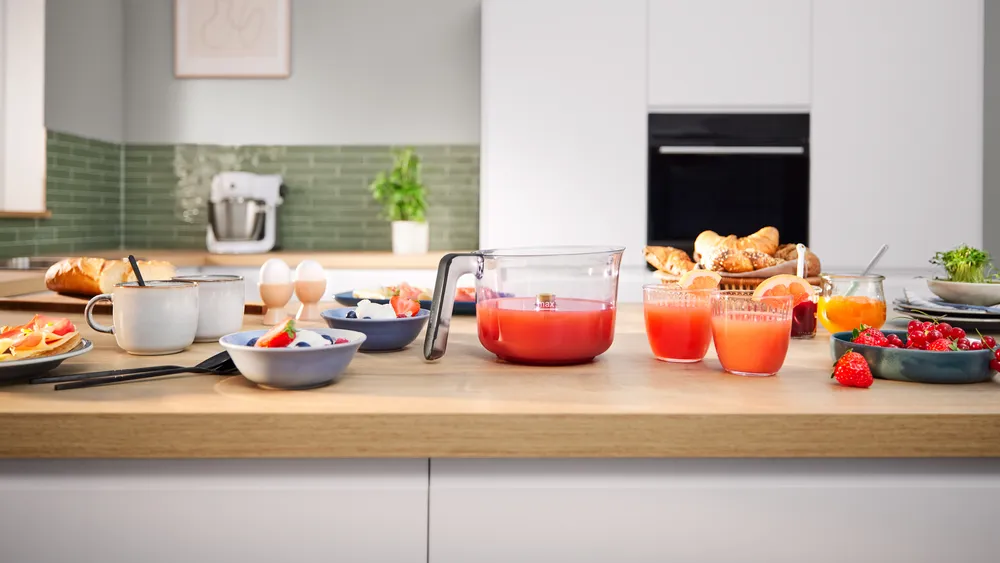 Egy reggelizőasztal ételekkel és italokkal: lé, kenyér, gyümölcs, tojás és croissant egy konyhában.