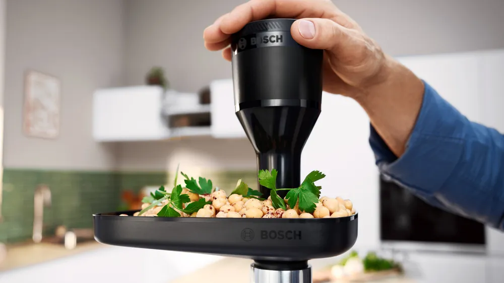 Egy kéz egy fekete Bosch konyhai robotgépet tart csicseriborsóval és petrezselyemmel teli tálca felett, konyhai környezetben.