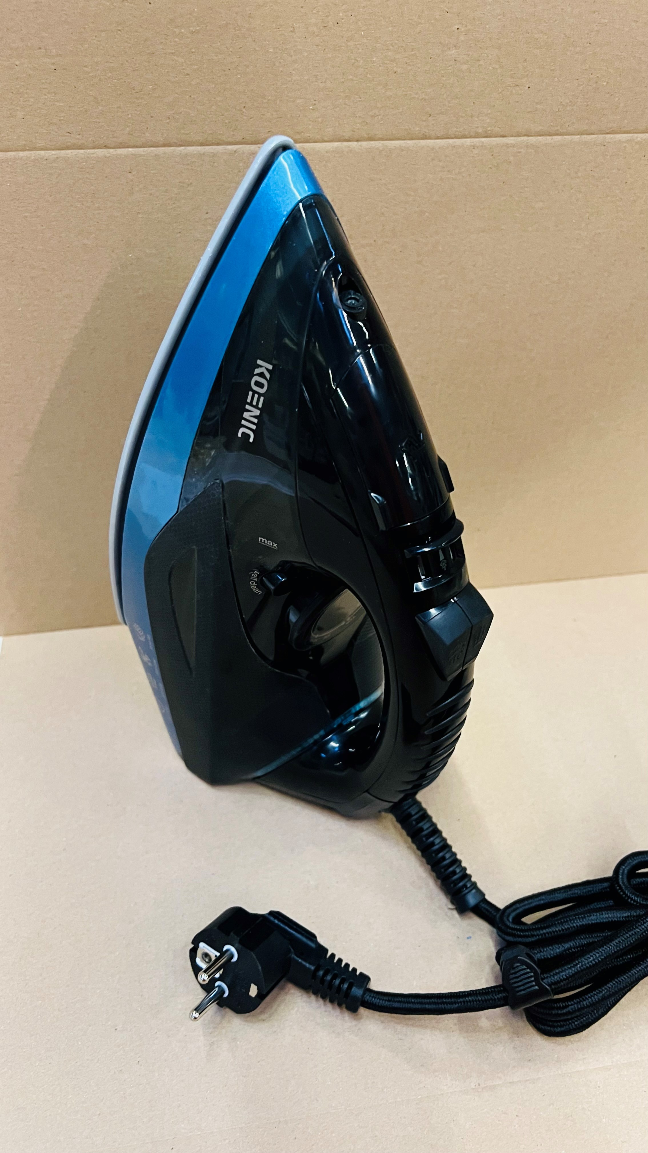 Czarno-niebieskie żelazko z czarnym kablem i europejską wtyczką. Leży na kartonowej powierzchni.