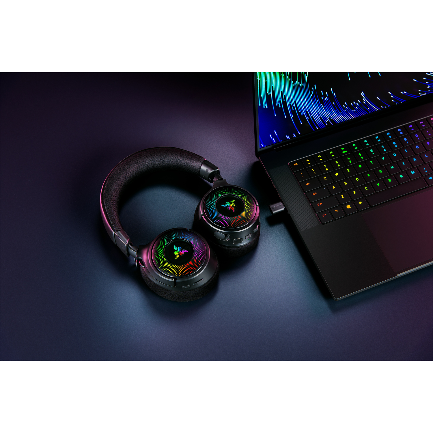 Hoofdtelefoon naast een laptop. De hoofdtelefoon gloeit met verschillende kleuren. Beide liggen op een donker oppervlak.