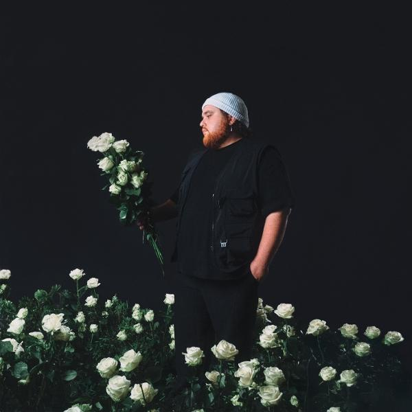 Ein Mann in Schwarz hält weiße Rosen in einem Rosenfeld, schwarzer Hintergrund.