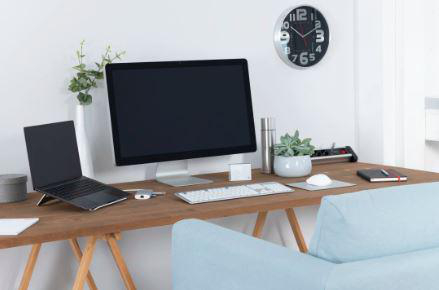 Home-Office-Setup. Laptop und Monitor auf einem Holzschreibtisch. Uhr an der Wand. Blaues Sofa und Pflanzen.