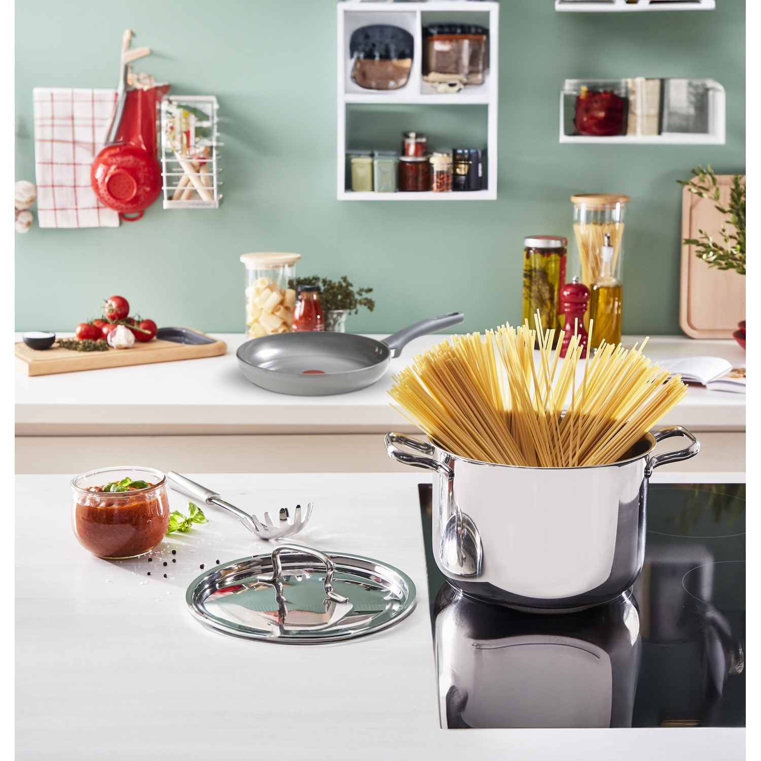 Scena di cucina con una padella, una pentola di pasta e ingredienti sul bancone e scaffali sullo sfondo.