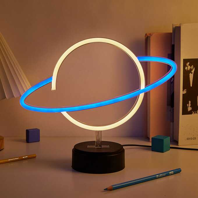 Lampa w kształcie planety z białym pierścieniem i niebieskim zewnętrznym. Stoi na czarnej podstawie. Kolorowe przedmioty.