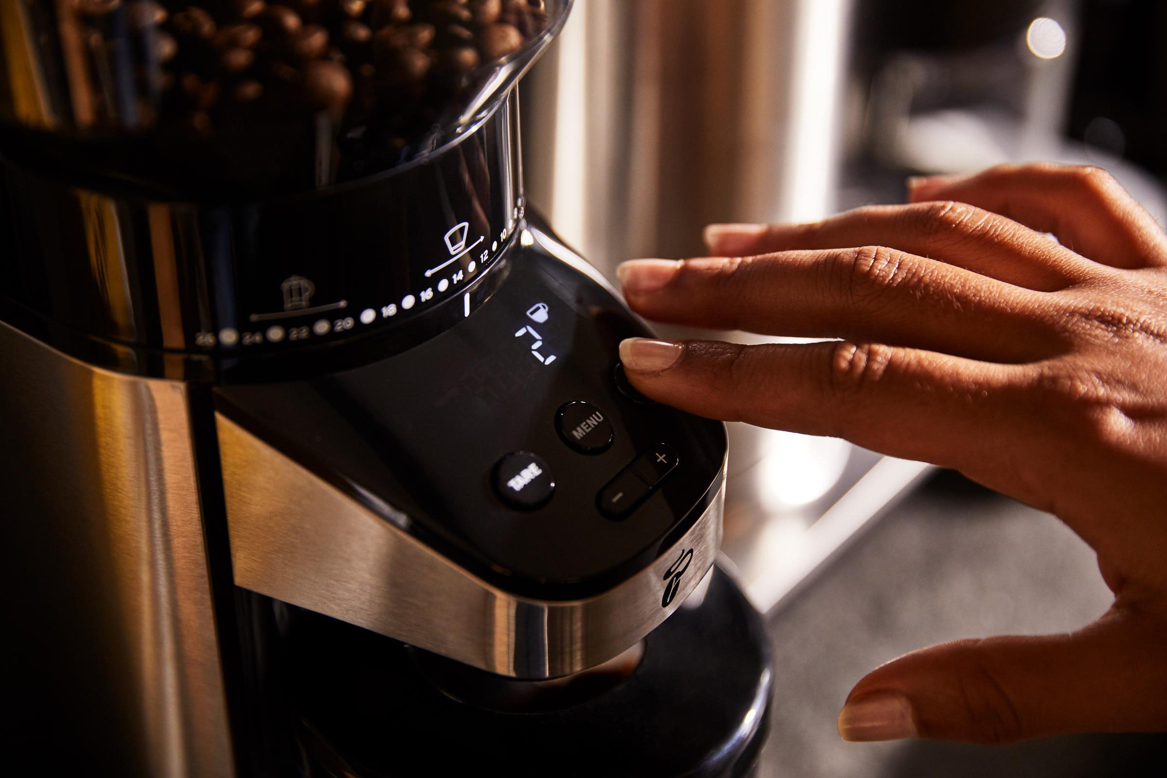 Eine Hand drückt eine Taste an einer schwarz-silbernen Kaffeemühle. Kaffeebohnen füllen die Oberseite.
