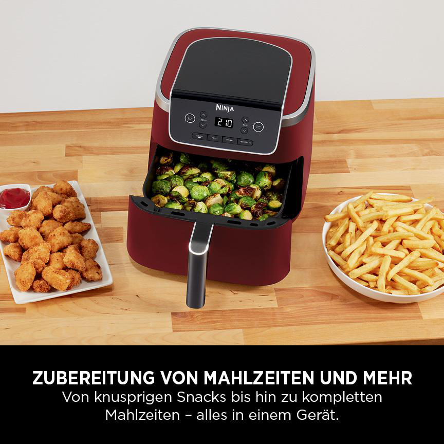 Rote Heißluftfritteuse mit Essen darin und Beilagen auf einer Holzoberfläche.