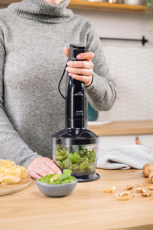 Személy bazsalikomot kever fekete turmixgéppel. Pesto egy tálban, tészta és dió egy fa felületen. Szürke pulóver.