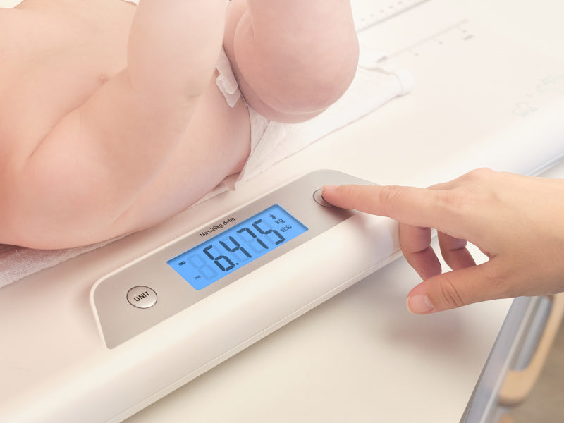 Baba a mérlegen. Egy kéz megnyom egy gombot, amely 6,475 kg-ot mutat. A baba a hasán fekszik.