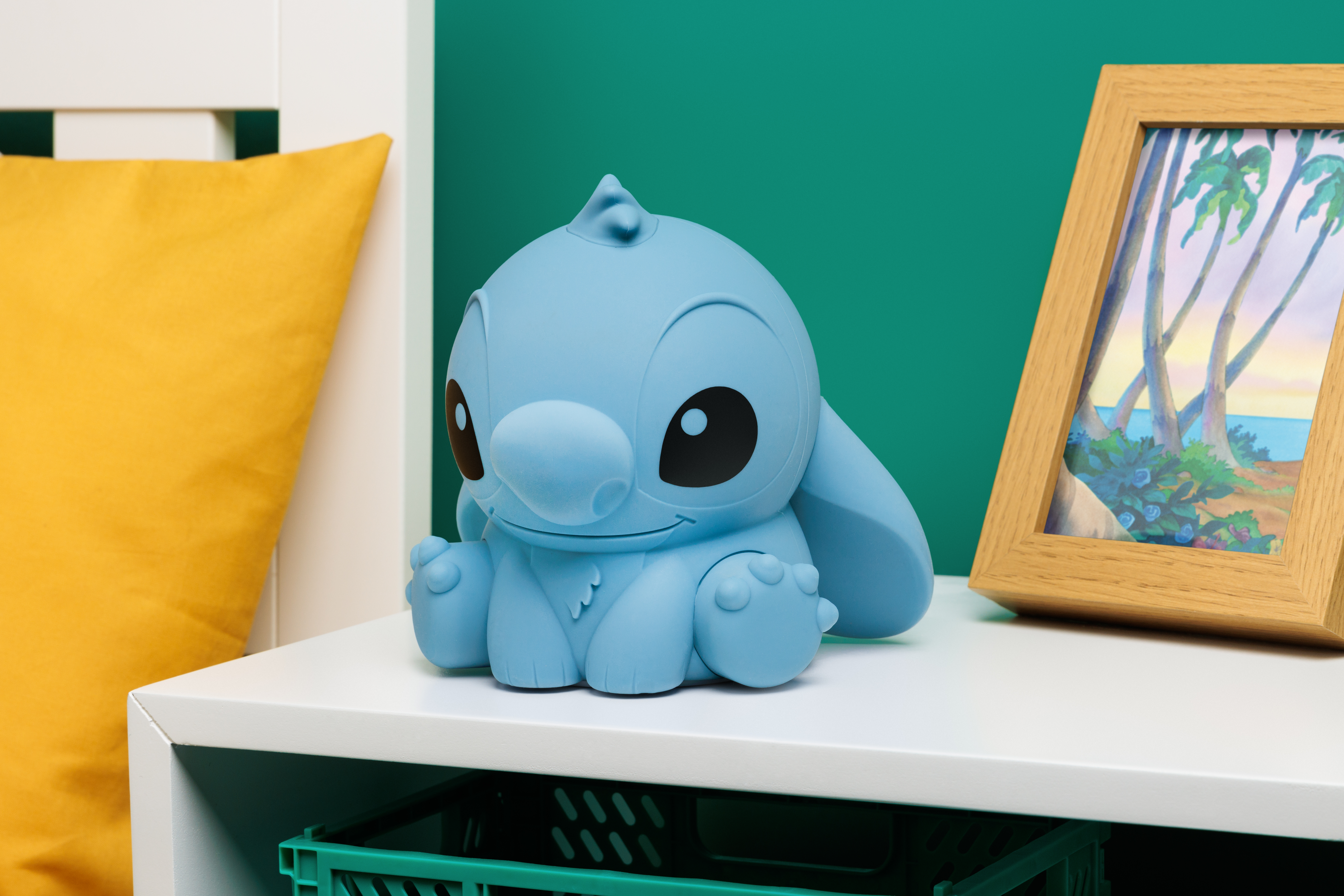 Eine Stitch-Figur sitzt auf einem weißen Tisch, mit einer grünen Kiste und einem gerahmten Bild.