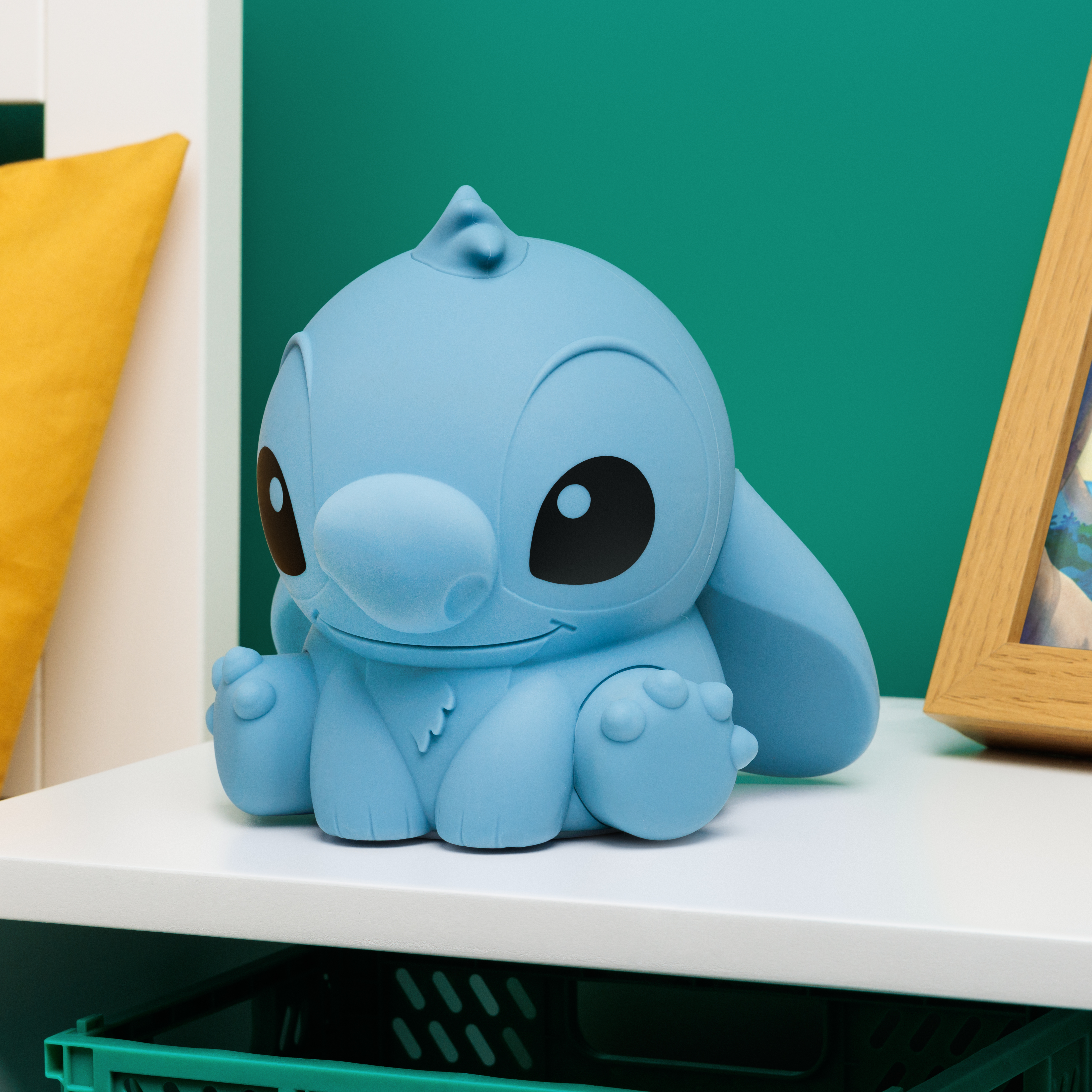 Blaue Stitch-Figur auf weißer Oberfläche. Ein gerahmtes Bild und eine grüne Box im Hintergrund.