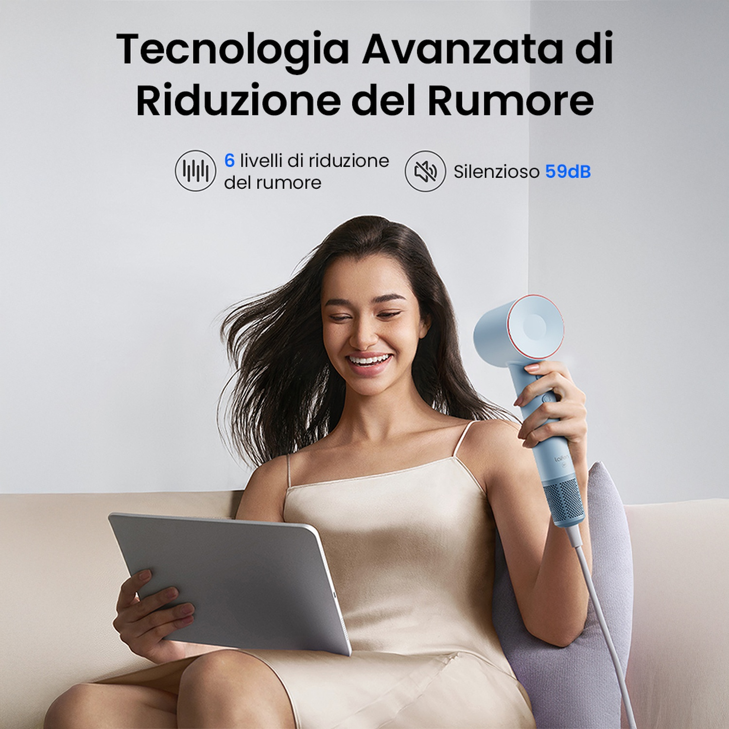 Una donna che tiene un asciugacapelli, sorridendo, con un tablet e un cuscino sul divano.