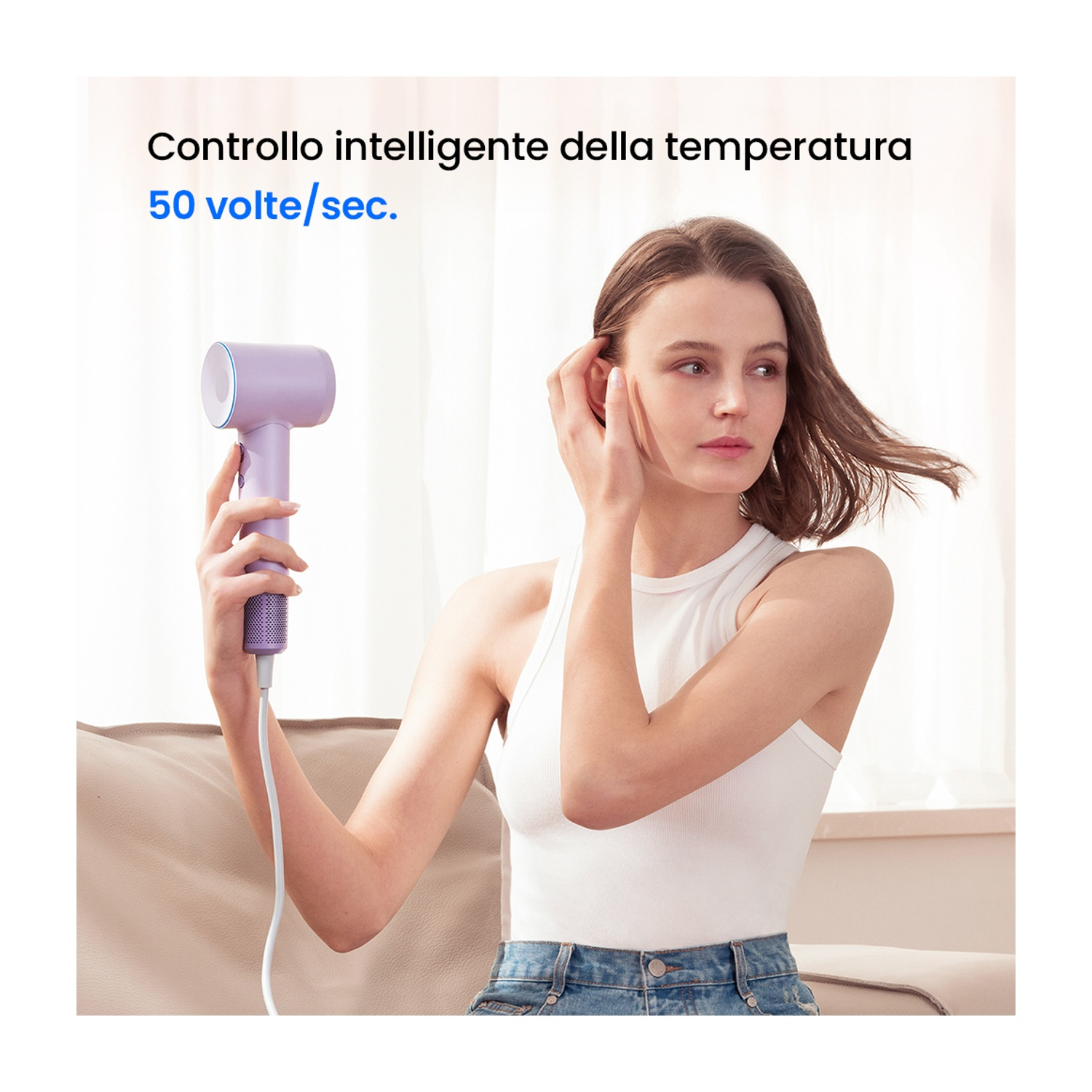 Una donna che si asciuga i capelli con un asciugacapelli viola. Indossa un top bianco ed è seduta su un divano.