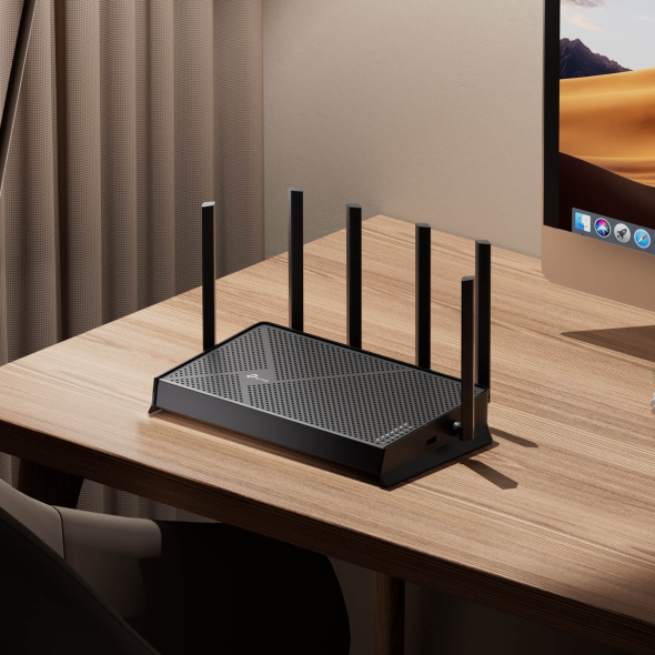 Fekete wifi router antennákkal egy fa asztalon, mögötte egy monitorral.