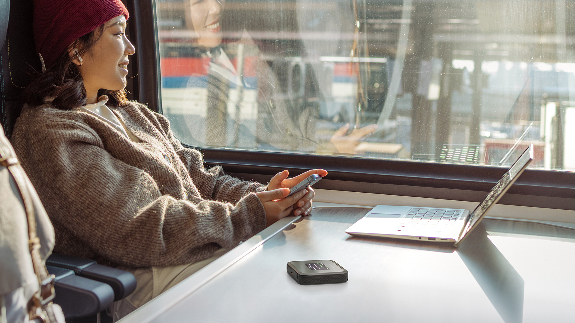 Frau im Zug, benutzt Handy und Laptop. Ein kleines Gerät liegt auf dem Tisch neben dem Laptop.