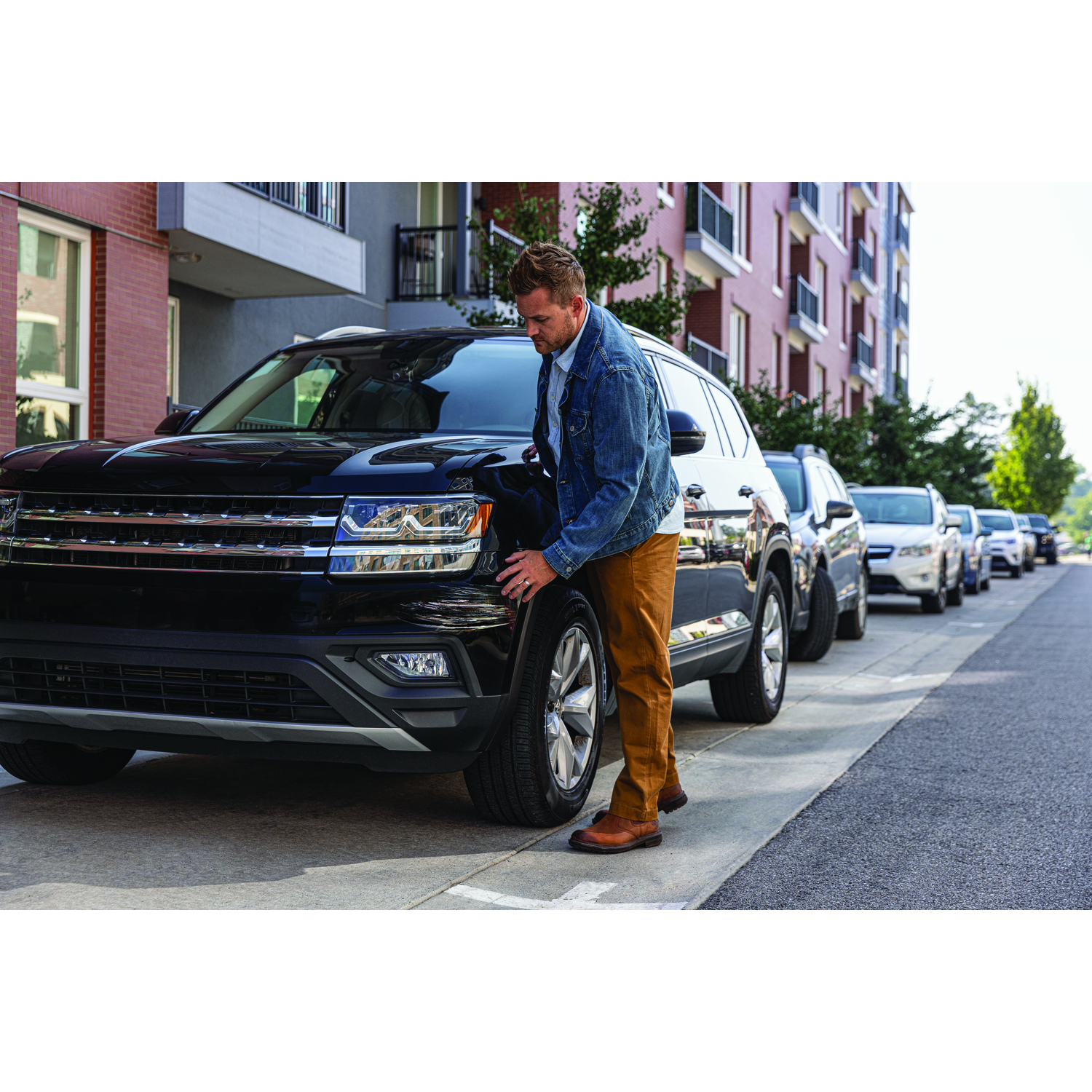 Een man bekijkt een zwarte SUV die op straat geparkeerd staat. Andere voertuigen en gebouwen zijn aanwezig.