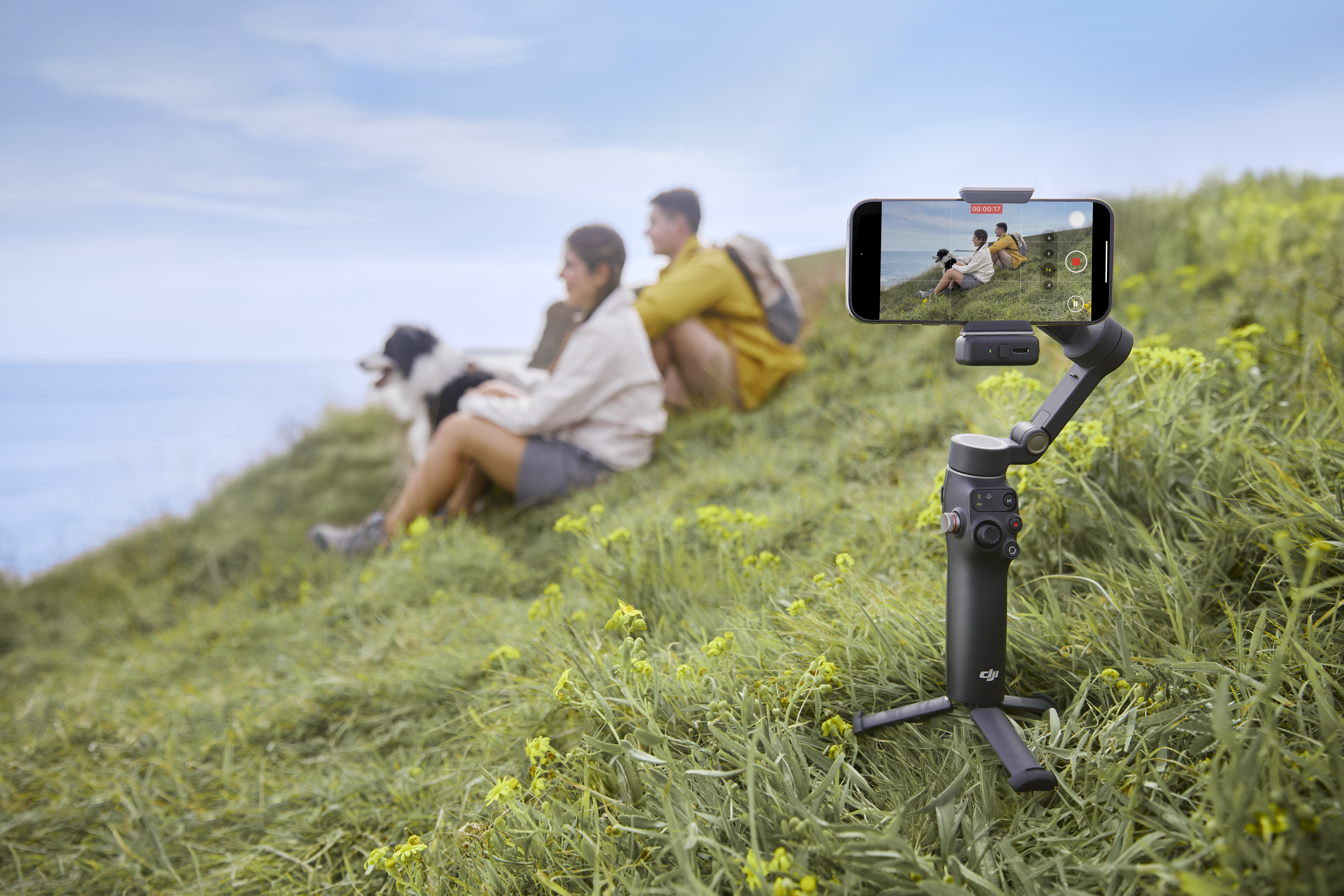 Leute filmen mit einem Telefon auf einem Stativ mit Blick auf das Meer. Grünes Gras und blauer Himmel.