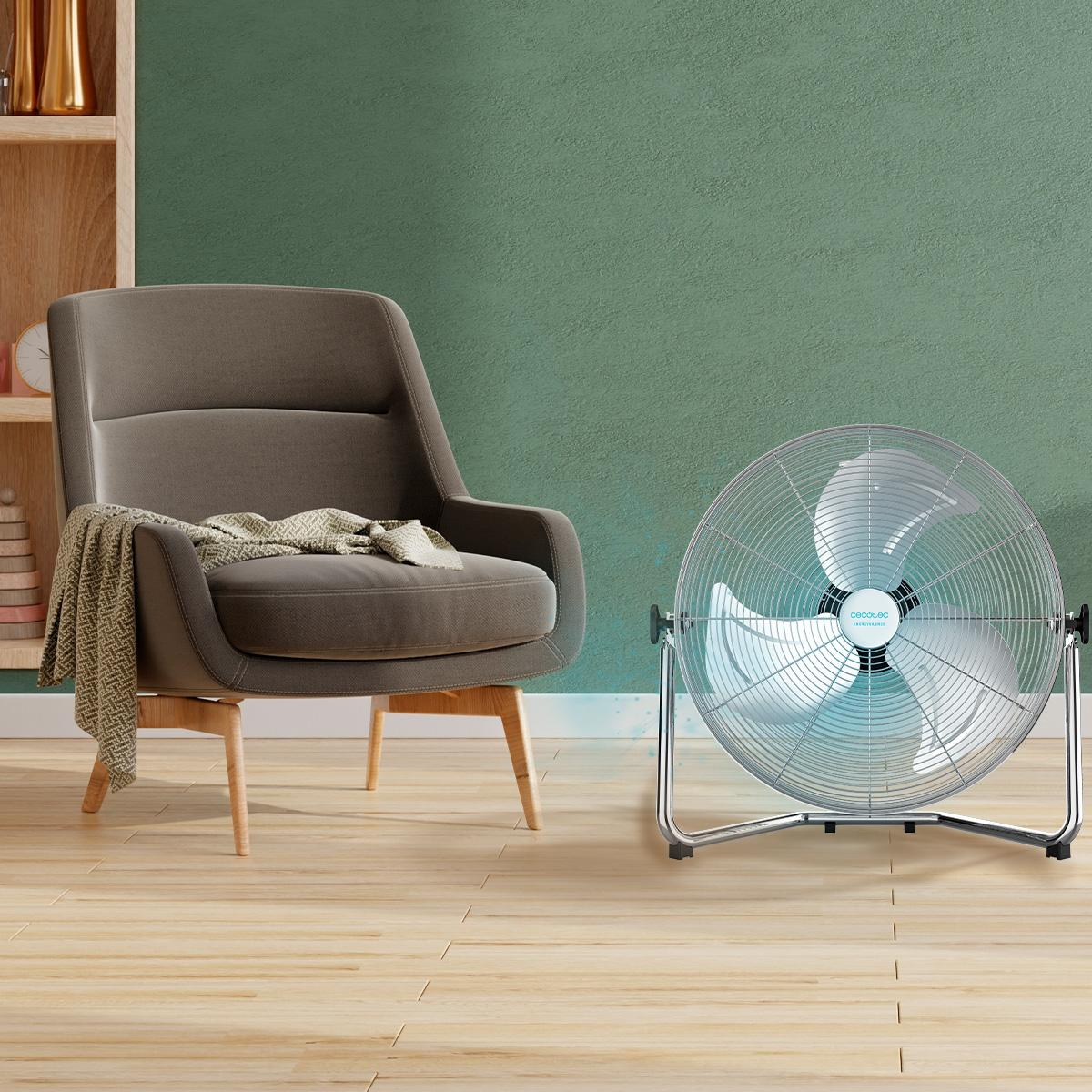 Un ventilador plateado frente a un sillón gris, con una pared verde en el fondo.