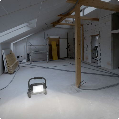 Innenraum einer Baustelle mit Scheinwerfer. Freiliegende Balken, unfertige Wände und Betonboden.