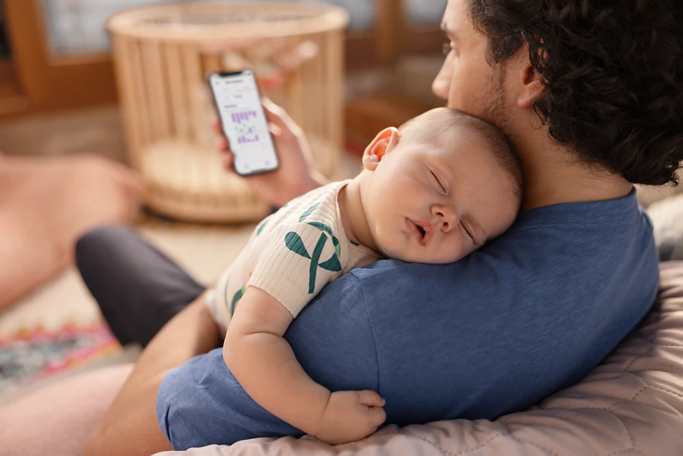 Egy férfi egy alvó babát tart a kezében. Telefonon a baba adatai láthatók.