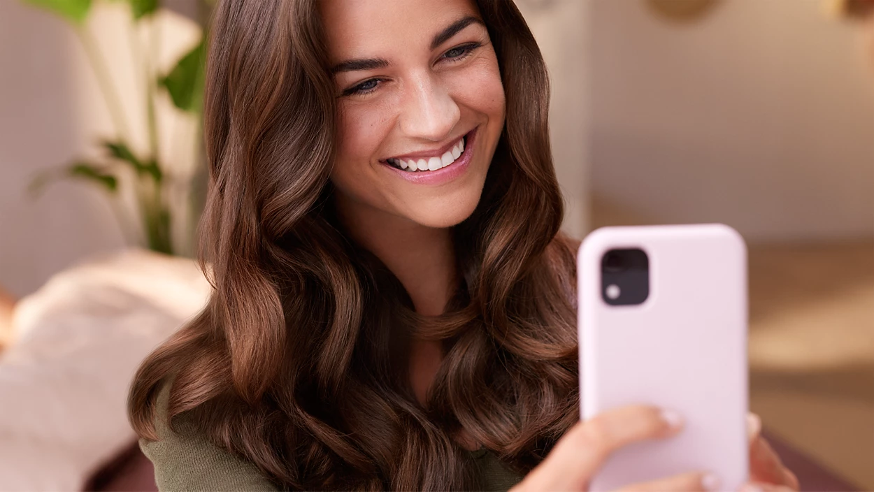 Nő mosolyog és a telefon képernyőjét nézi, göndör barna hajjal és világos rózsaszín telefontokkal.