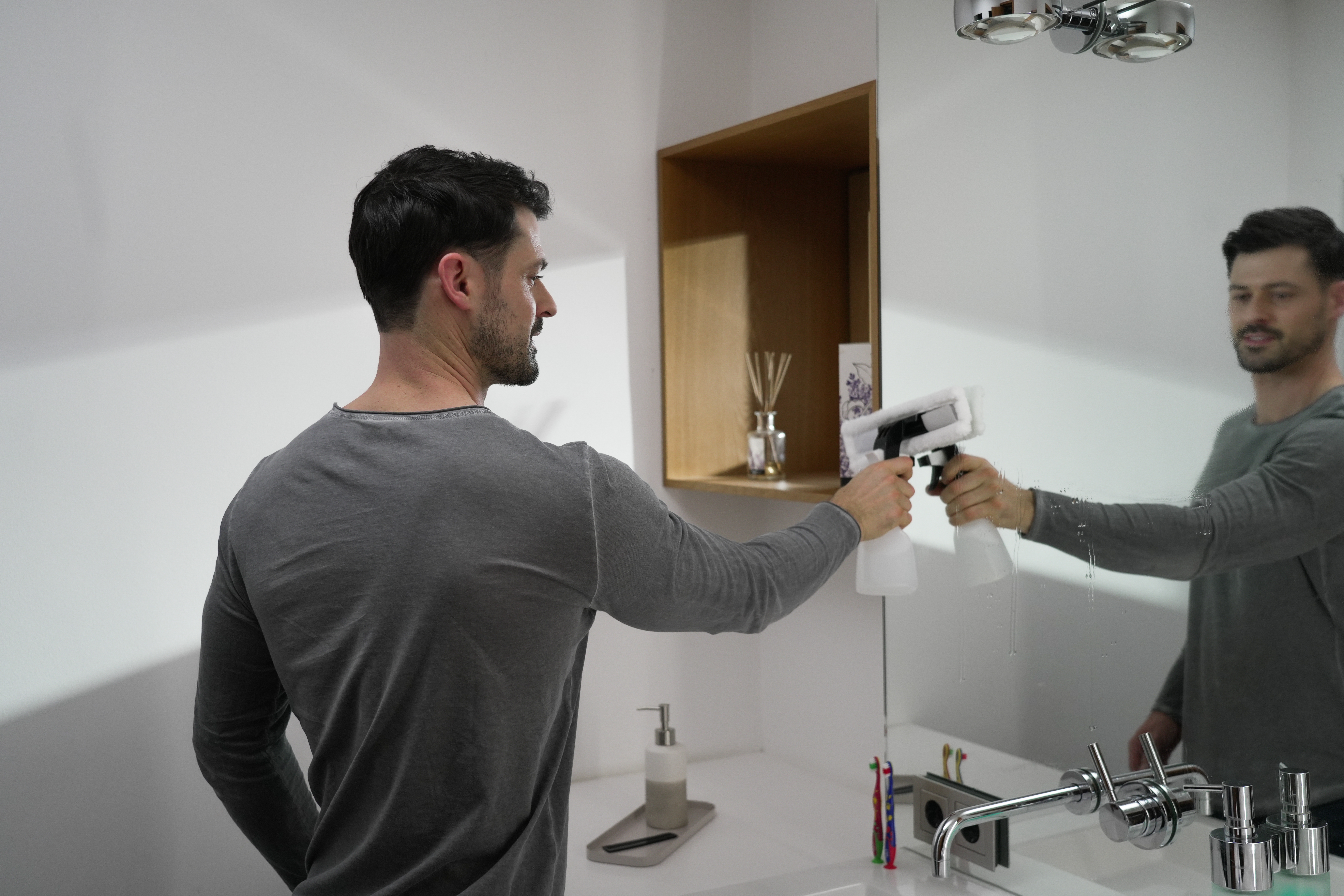 Mann sprüht einen Spiegel mit einem Reinigungswerkzeug ein. Er steht in einem Badezimmer mit sauberer Arbeitsplatte.