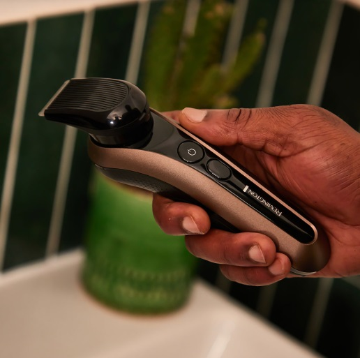 Dłoń trzyma trymer do brody Remington. Jest złoty i czarny na tle sceny łazienkowej.