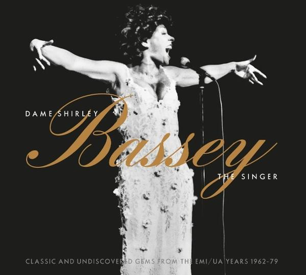 Dame Shirley Bassey mit offenen Armen, singt ins Mikrofon, auf dunklem Hintergrund.