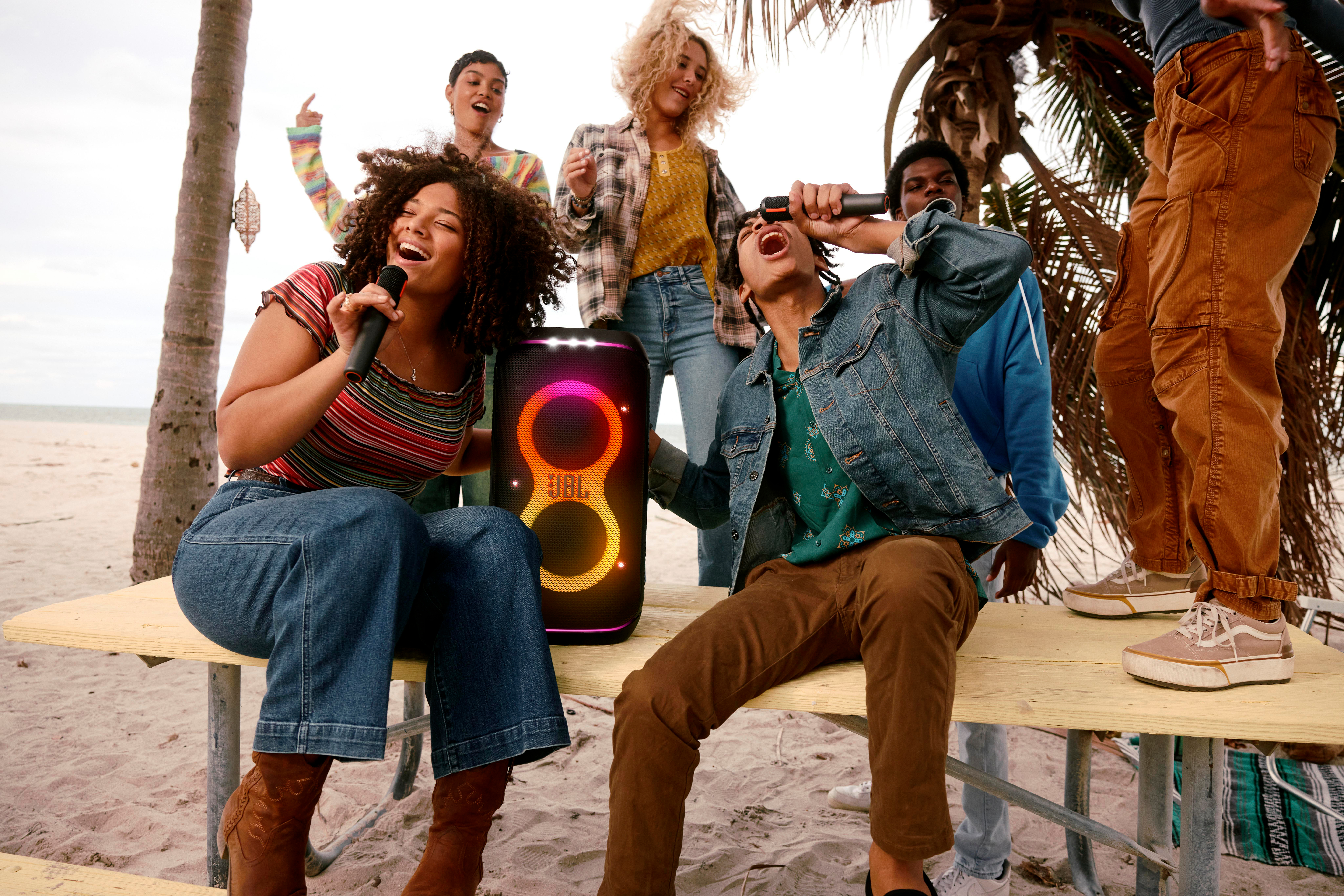 Eine Gruppe von Leuten, die mit einem JBL-Lautsprecher am Strand singen. Der Himmel ist bewölkt.