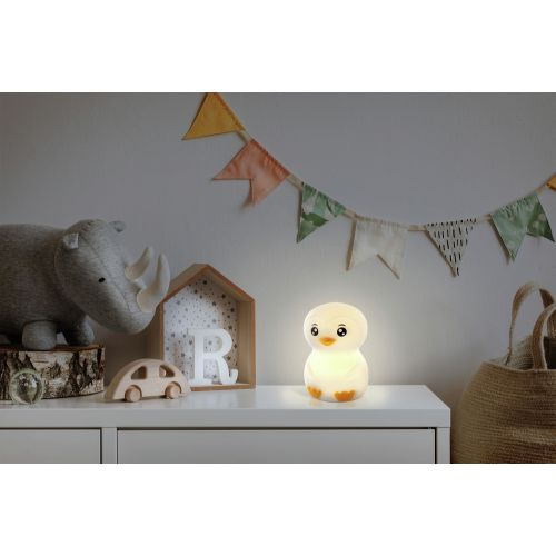 Eine weiße und gelbe Pinguinlampe auf einer weißen Kommode. Es gibt ein Spielzeugauto und ein Holzhaus.