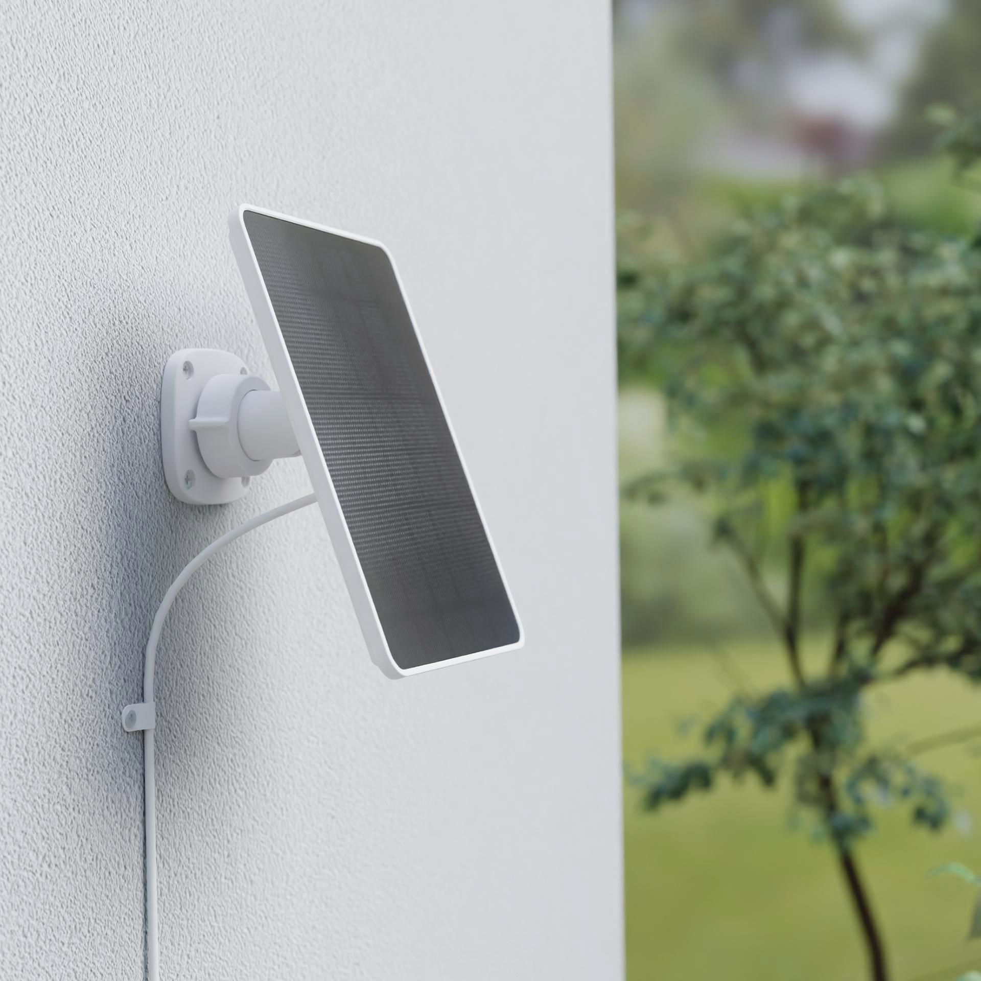 Weißes Solarpanel an grauer Wand mit Kabel. Grüne Bäume im Hintergrund.