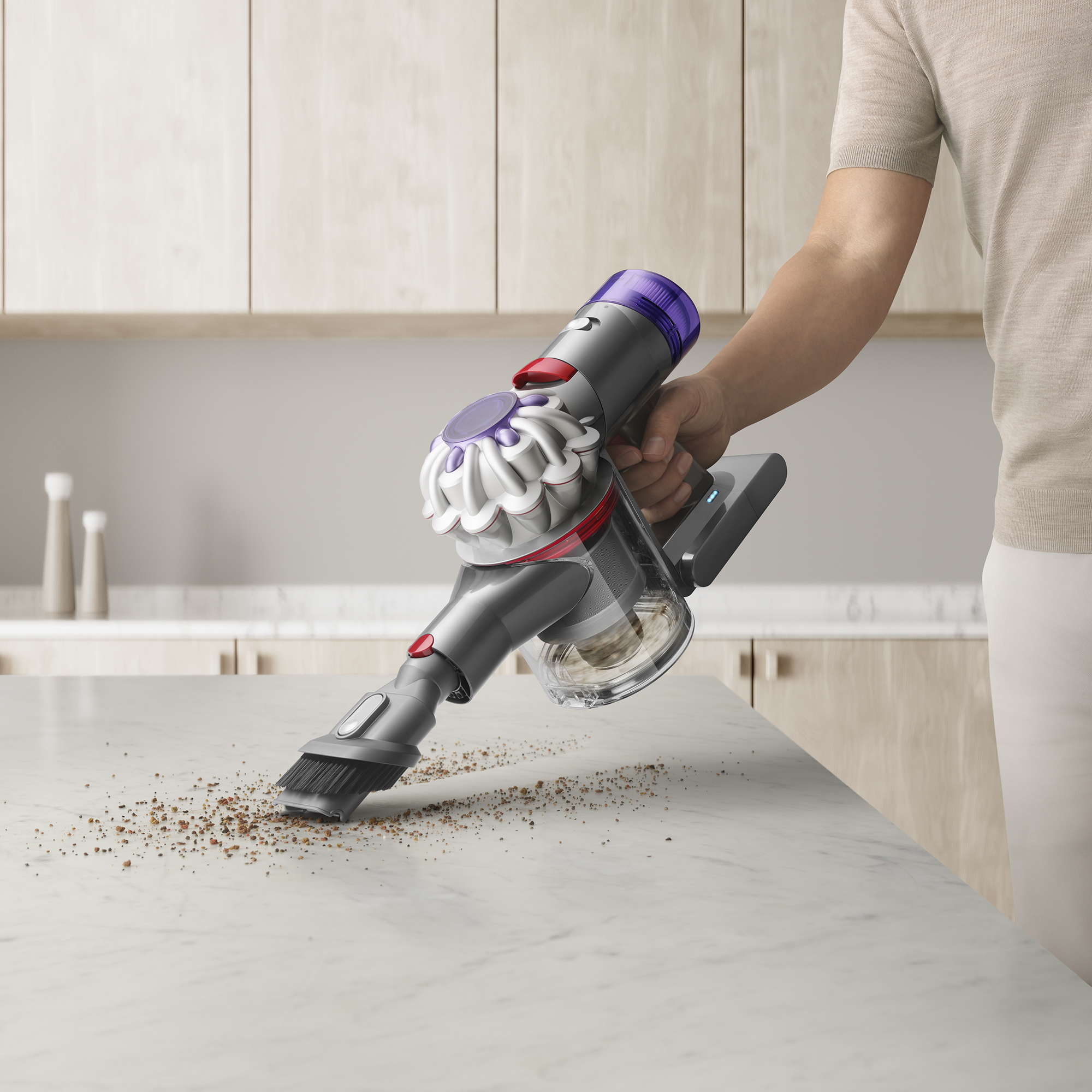 Eine Person reinigt eine weiße Arbeitsplatte mit einem grau-violetten Staubsauger. Holzschränke im Hintergrund.