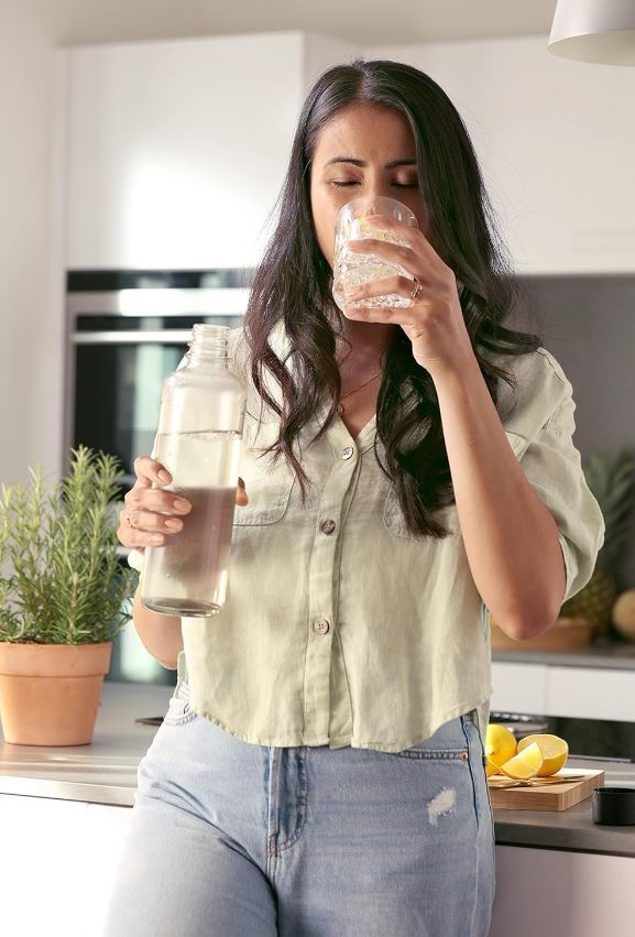 Frau in der Küche trinkt aus einem Glas. Sie hält eine Wasserflasche. Zitronen und Pflanze im Vordergrund.