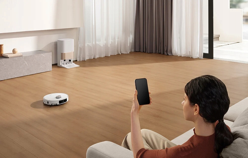 Nő telefonnal irányítja a robotporszívót egy modern nappaliban.