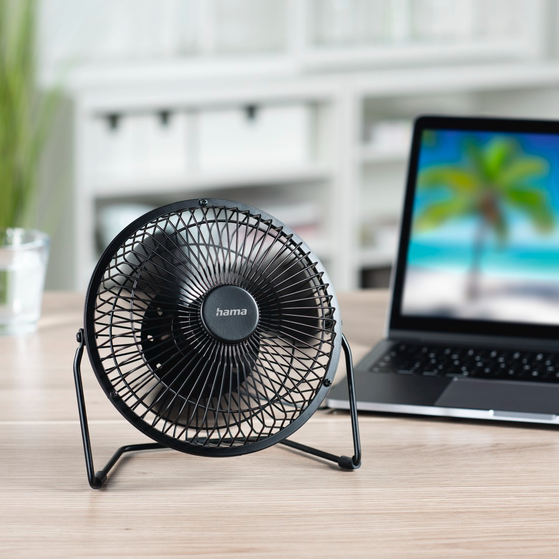 Fekete asztali ventilátor faasztalon. A háttérben egy laptop strandjelenettel.