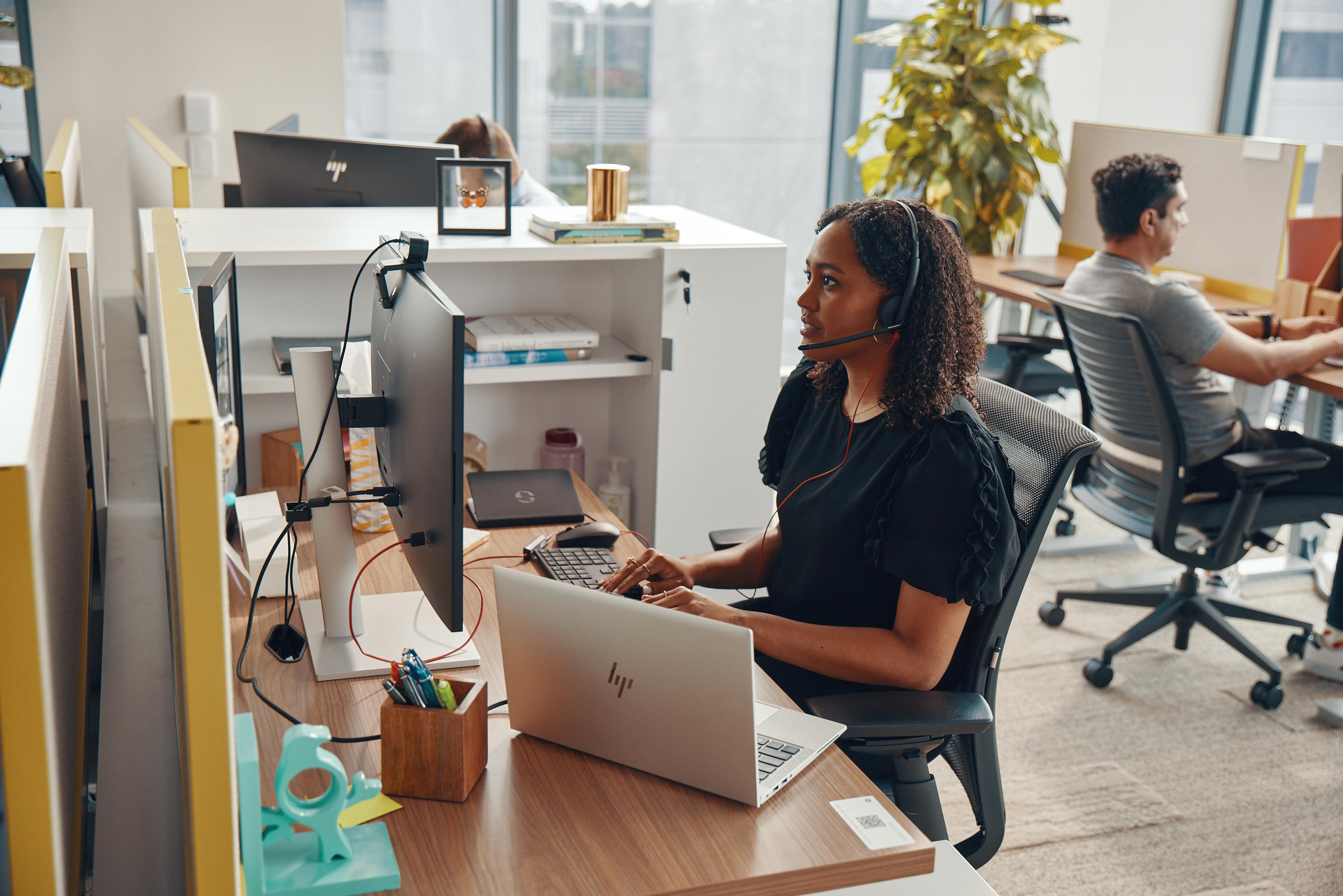 Frau mit Headset arbeitet an einem Schreibtisch im Büro, Laptop und Monitor sichtbar.