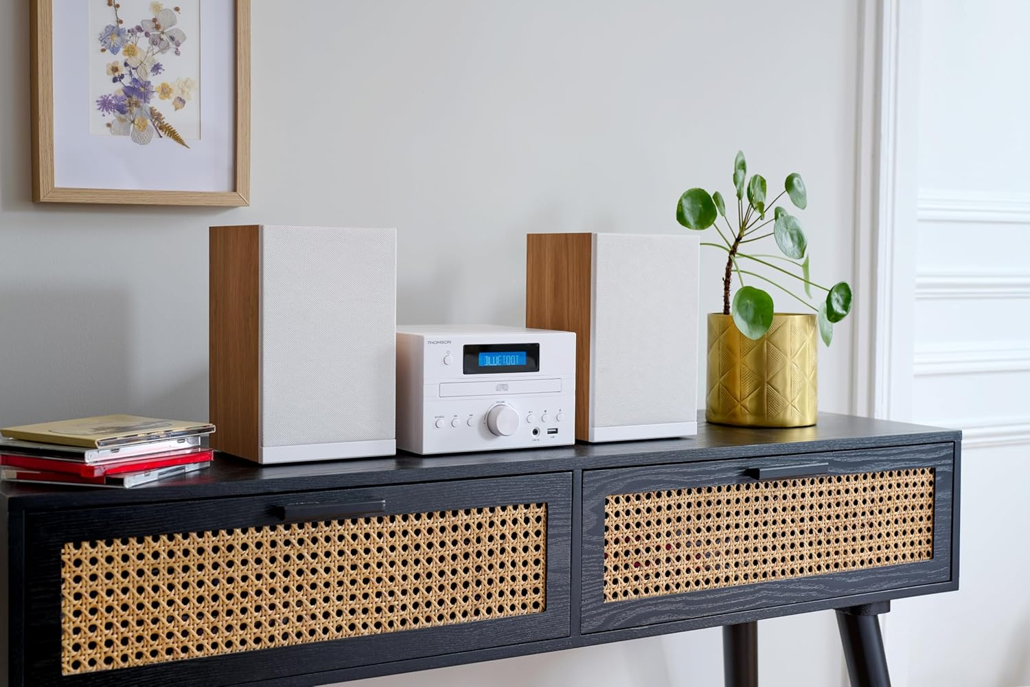 Sistema de audio blanco sobre un mueble negro, con altavoces. Una planta y arte enmarcado están en el mueble.