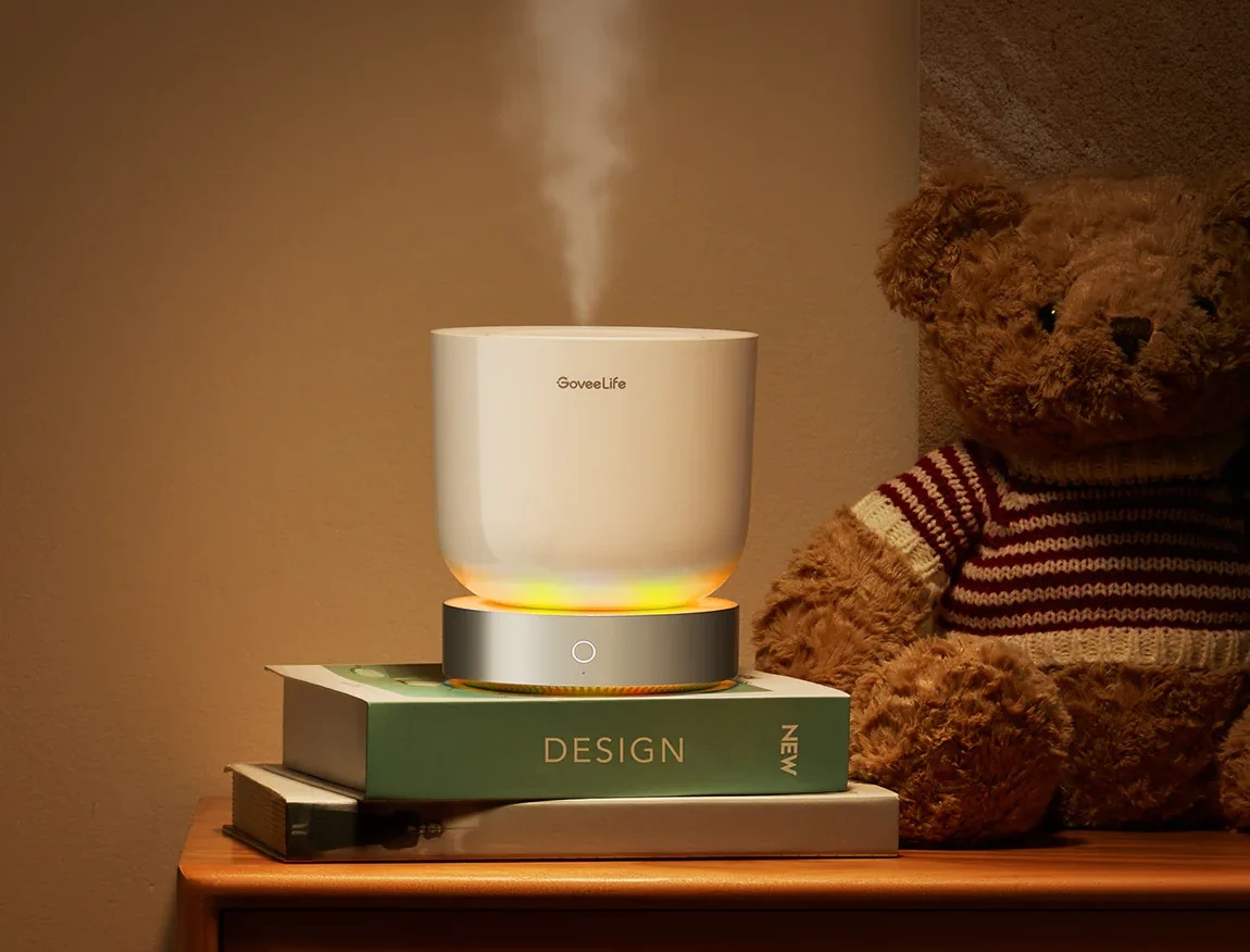 Biały nawilżacz na książkach, para unosi się, obok pluszowego misia. Ciepłe oświetlenie. Tło to ściana.