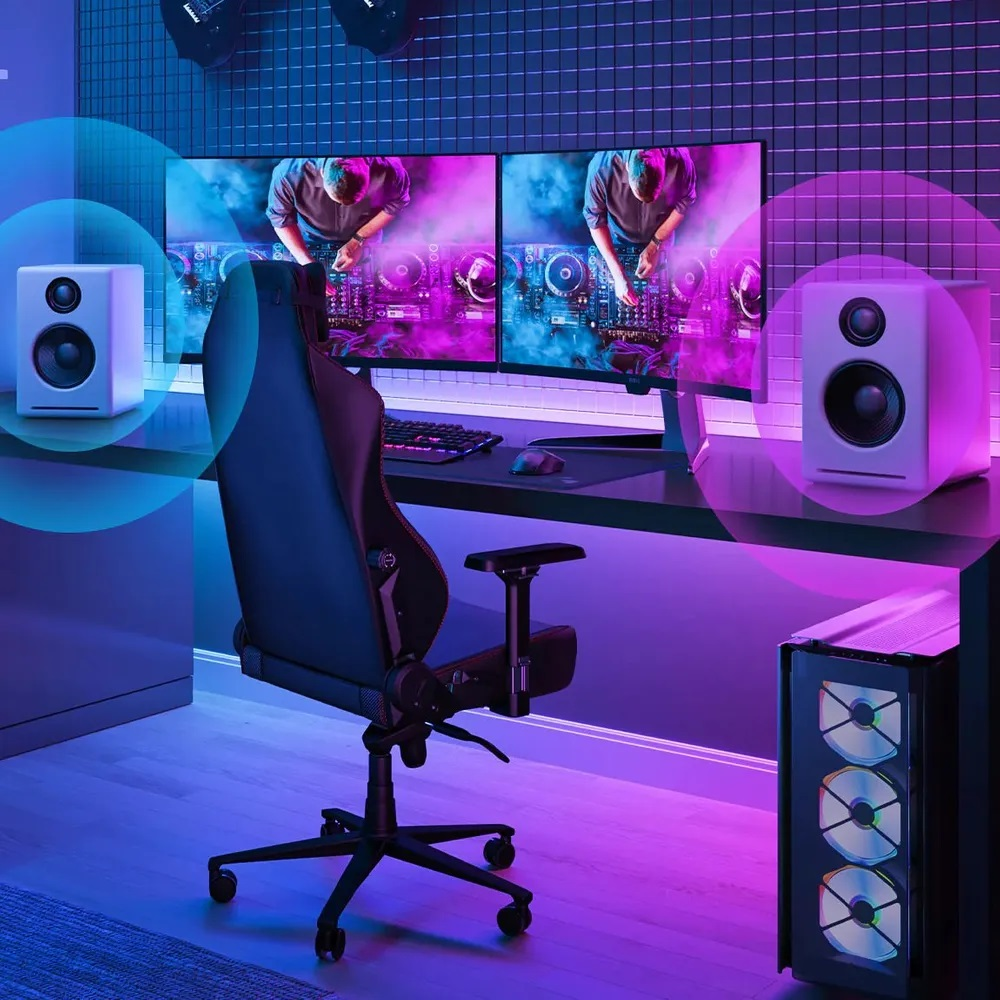 Konfiguracja do gier: krzesło, monitory, głośniki, PC i klawiatura. Scena jest oświetlona na niebiesko i fioletowo.