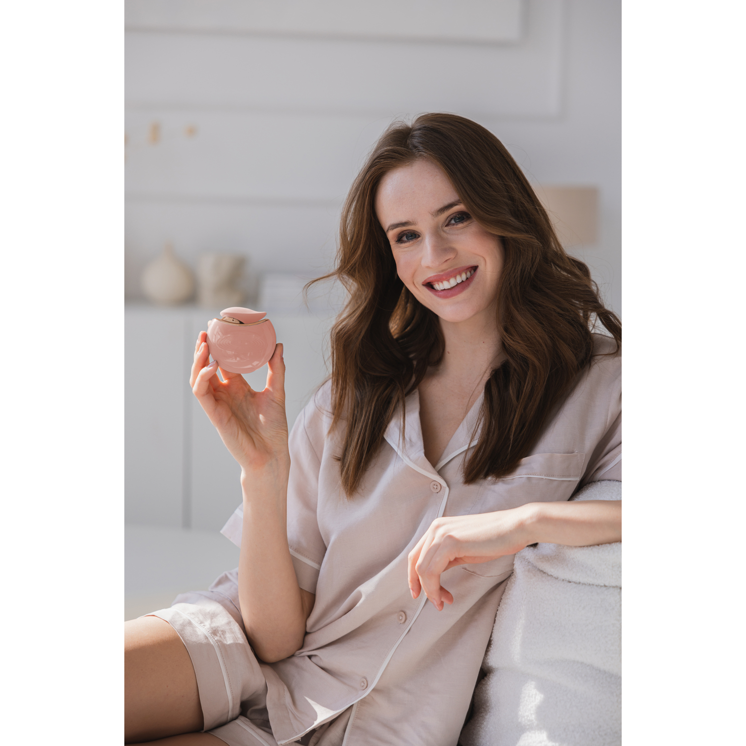A smiling woman in pajamas sits, holding a pink beauty device in a sunny room.
