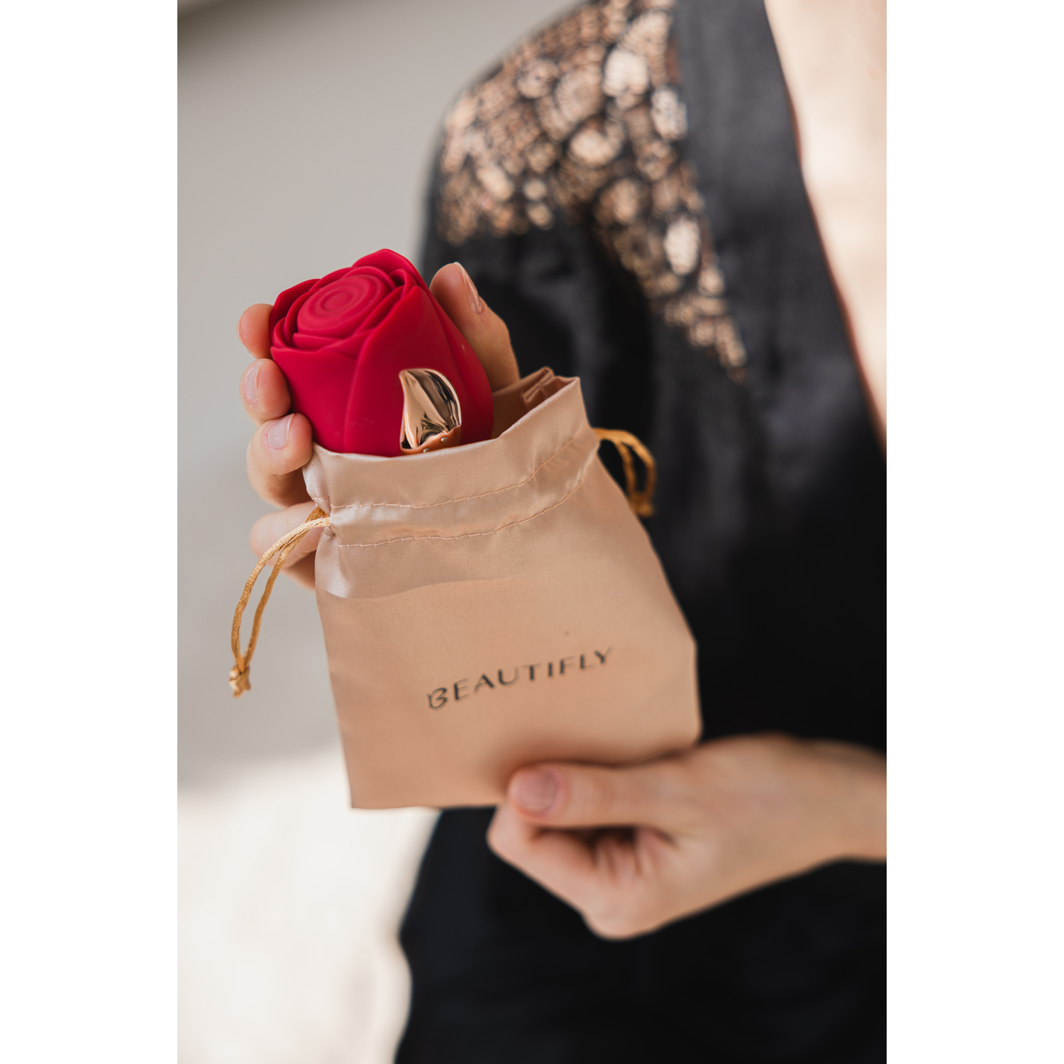 Hand holding a red rose device in a beige bag. The bag says BEAUTIFLY. Black robe background.