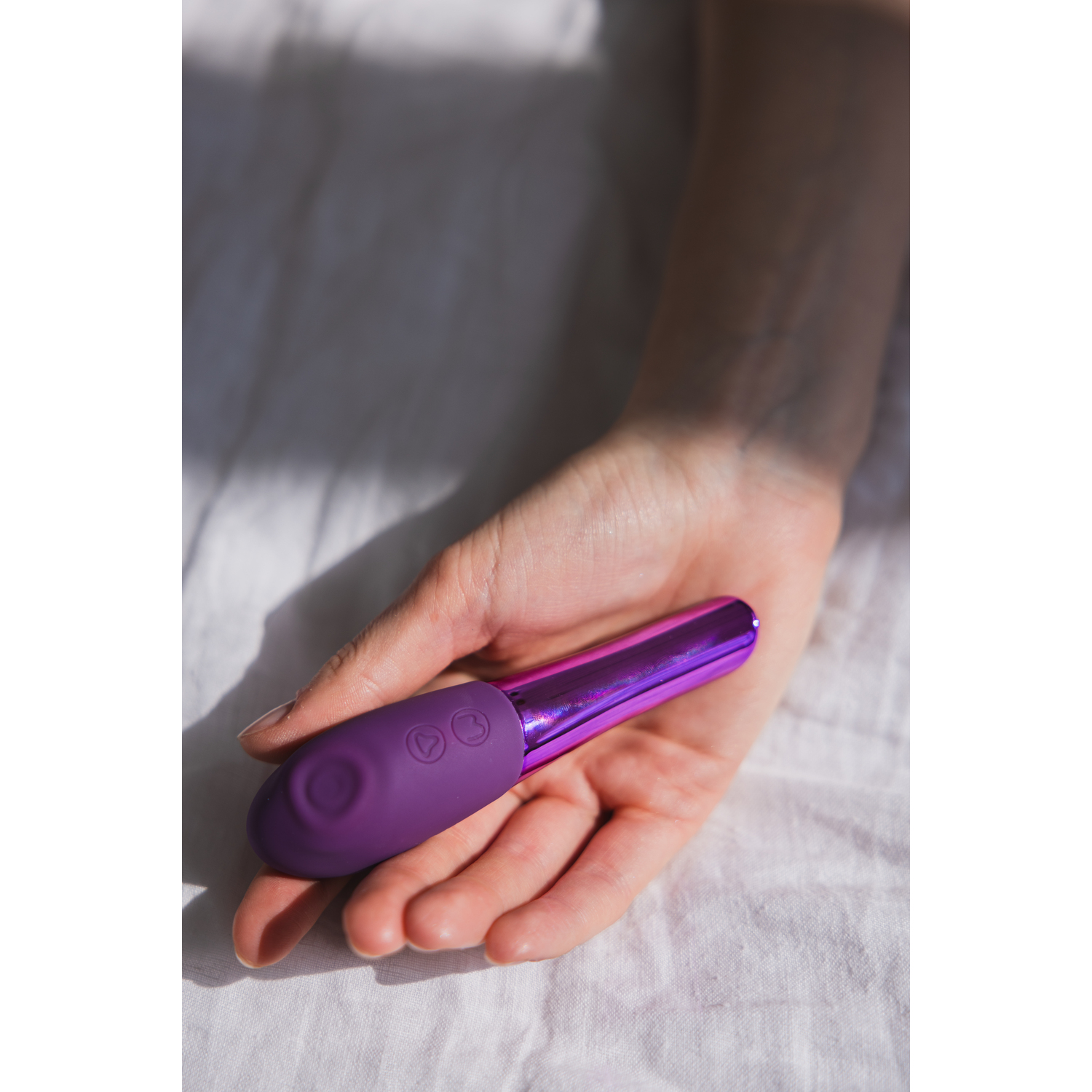 Hand holding a purple vibrator, set on a white bedsheet, lit by sunlight.