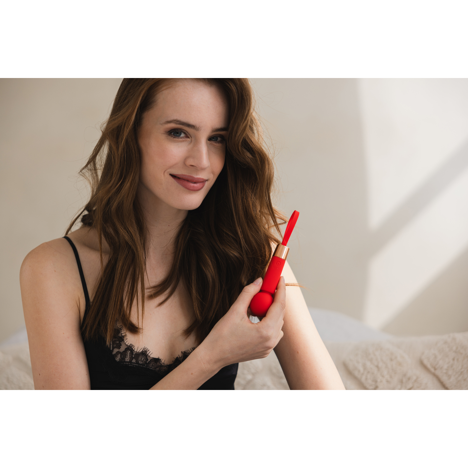 Woman holding a red sex toy, smiling, wearing black pajamas.